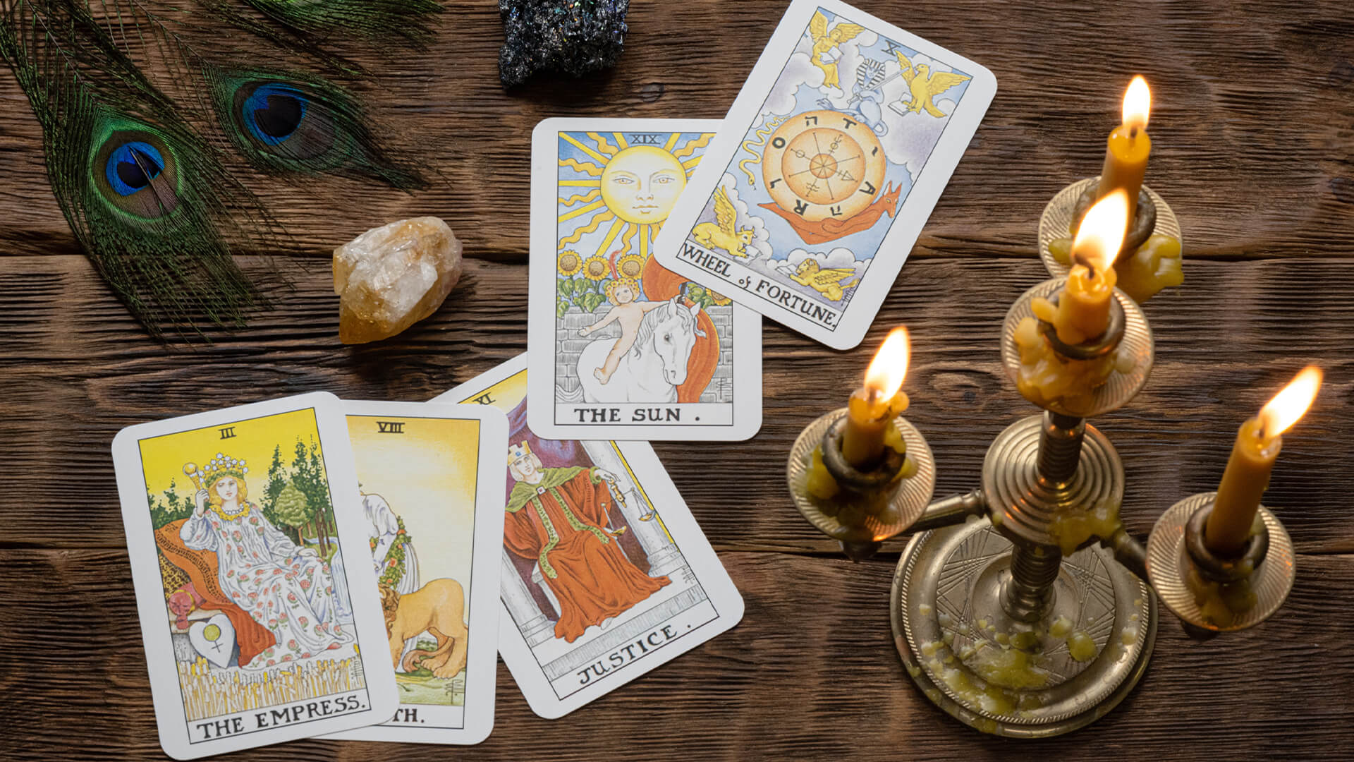 A selection of Tarot cards on a wooden table top, with peacock feathers and a candelabra