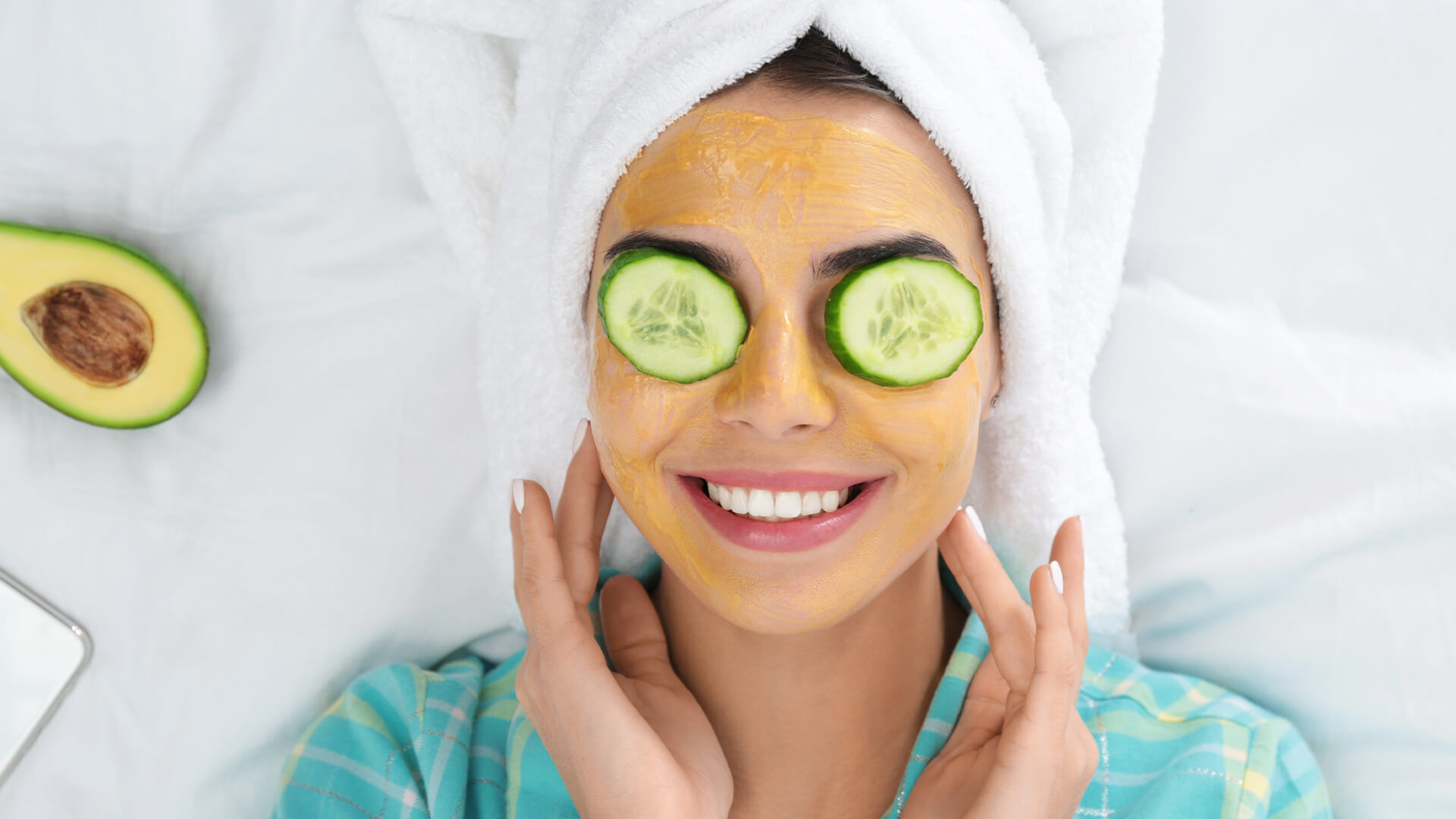 Woman with a facemask on, cucumbers on her eyes, and her hair wrapped in a towel