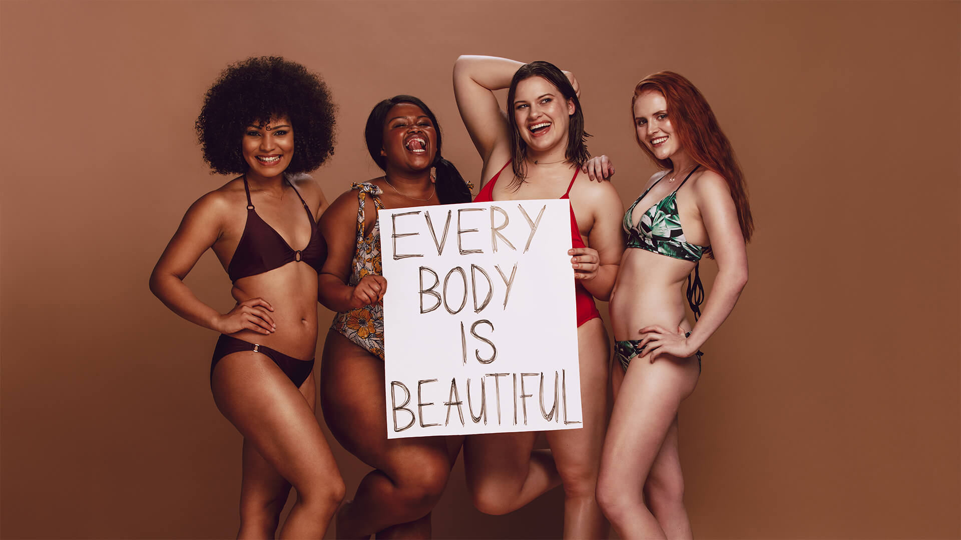 Four beautiful women of different shapes and sizes in bikinis, holding a sign that reads 