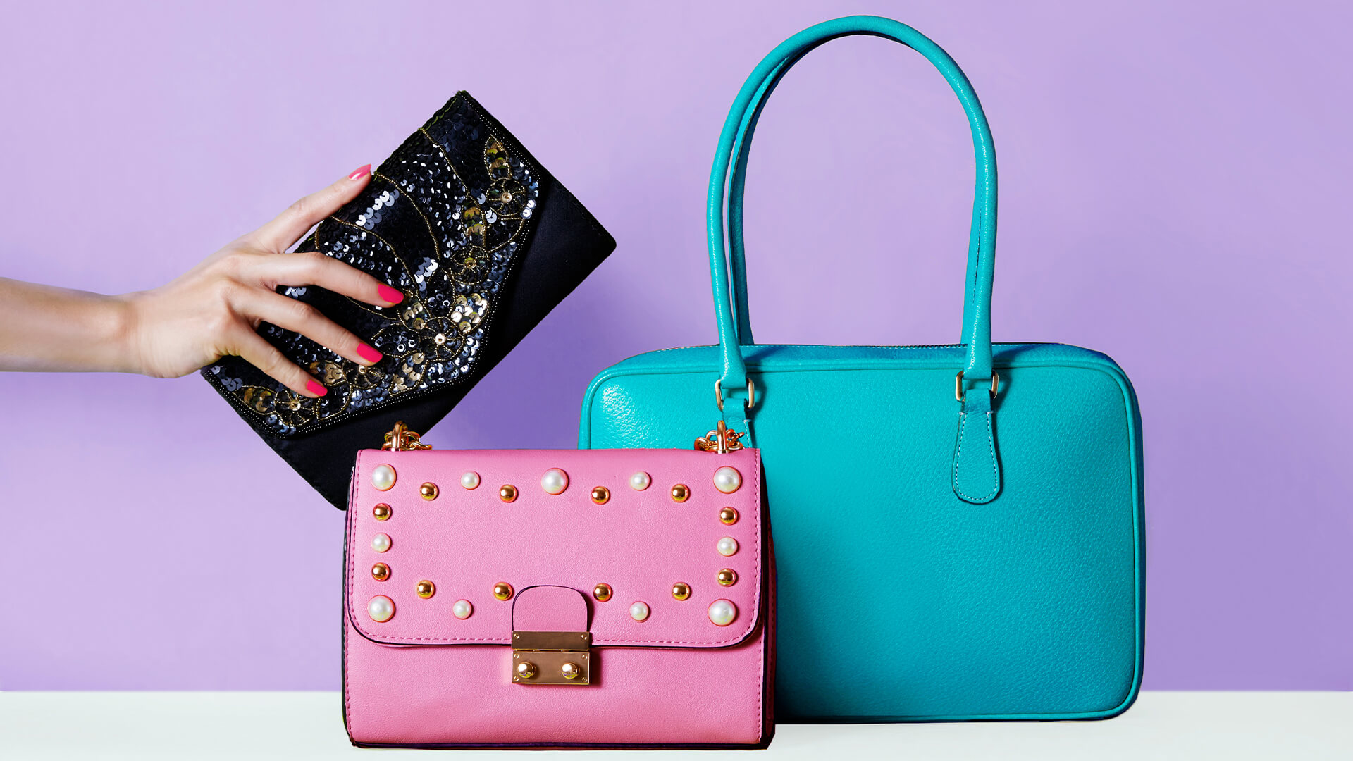 Three luxury bags against a lilac background. One is a larger turquois pocketbook, one is a bubblegum pink shoulder bag with pearl details, and the third is a patent black clutch