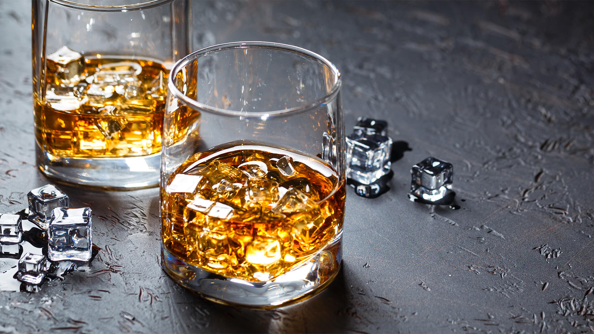 Two Whiskey glasses, on a slate surface. There is whiskey and ice in each glass, and a few scattered ice cubes on the surface