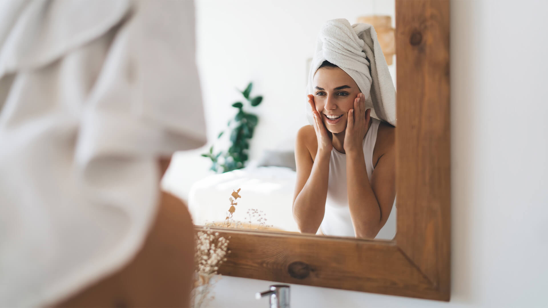 Young beautiful female with towel on head smiling to mirror reflection standing in the bathroom at home.