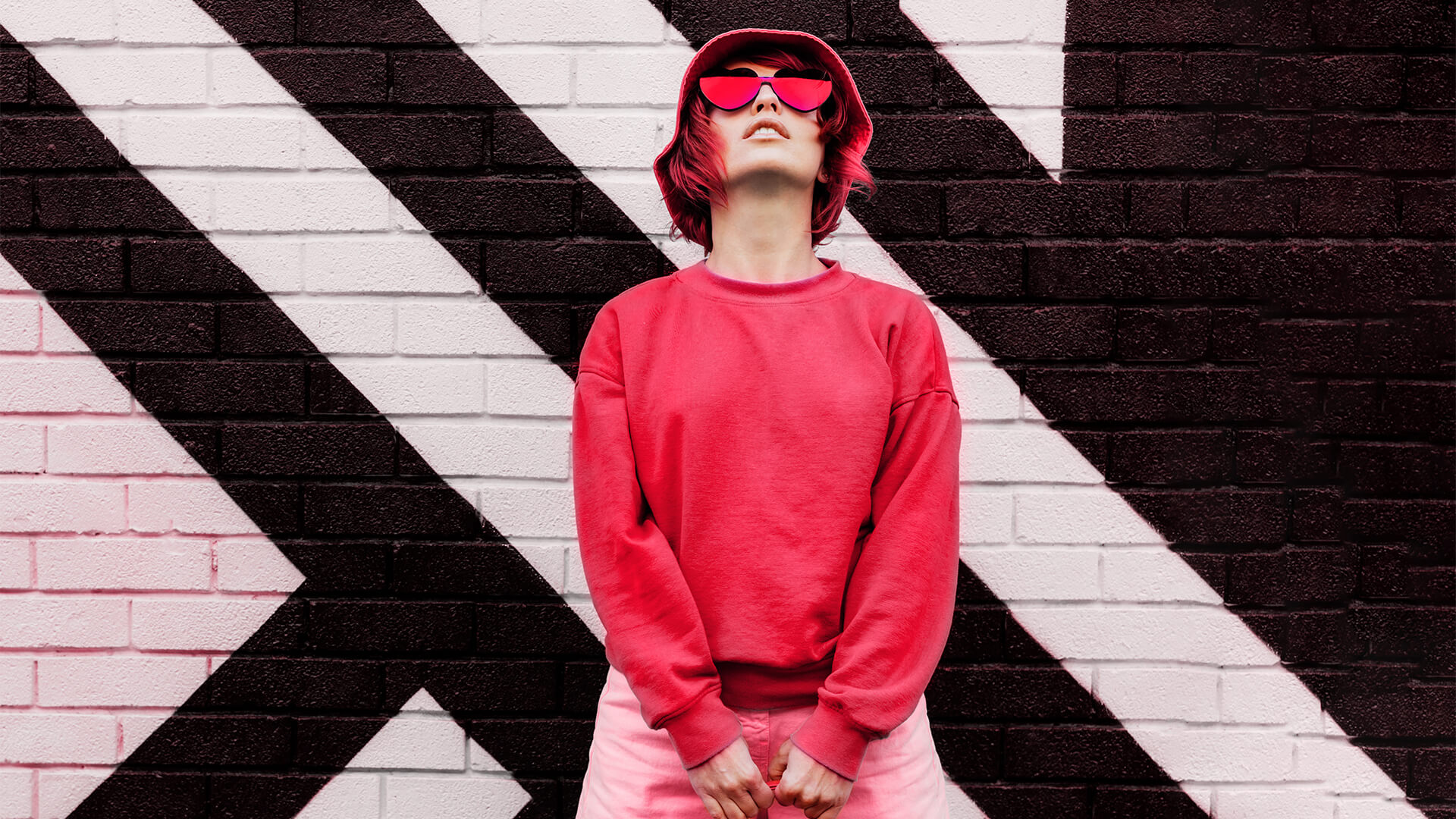 Hipster fashion young woman in trendy magenta color sweatshirt and sunglasses and bucket hat posing on the painted brick wall background.