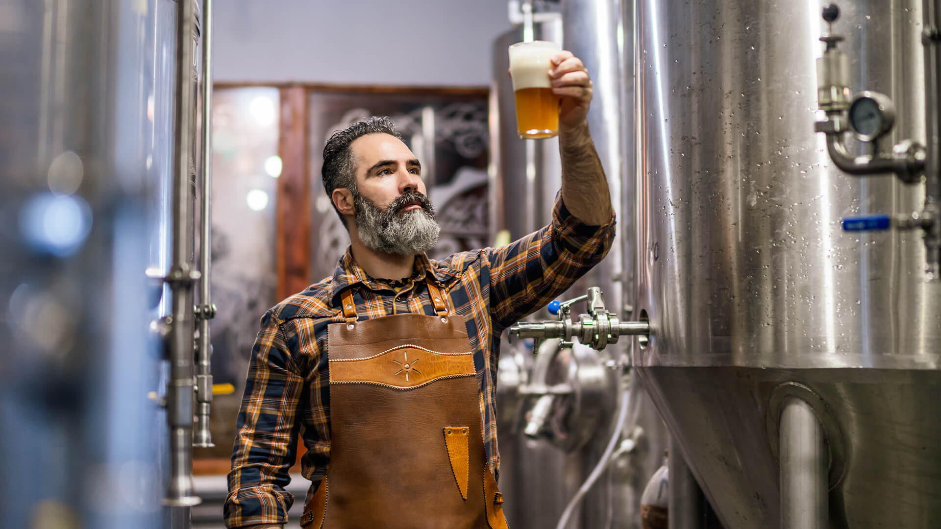 Bearded brewery master holding glass of beer and evaluating its visual characteristics