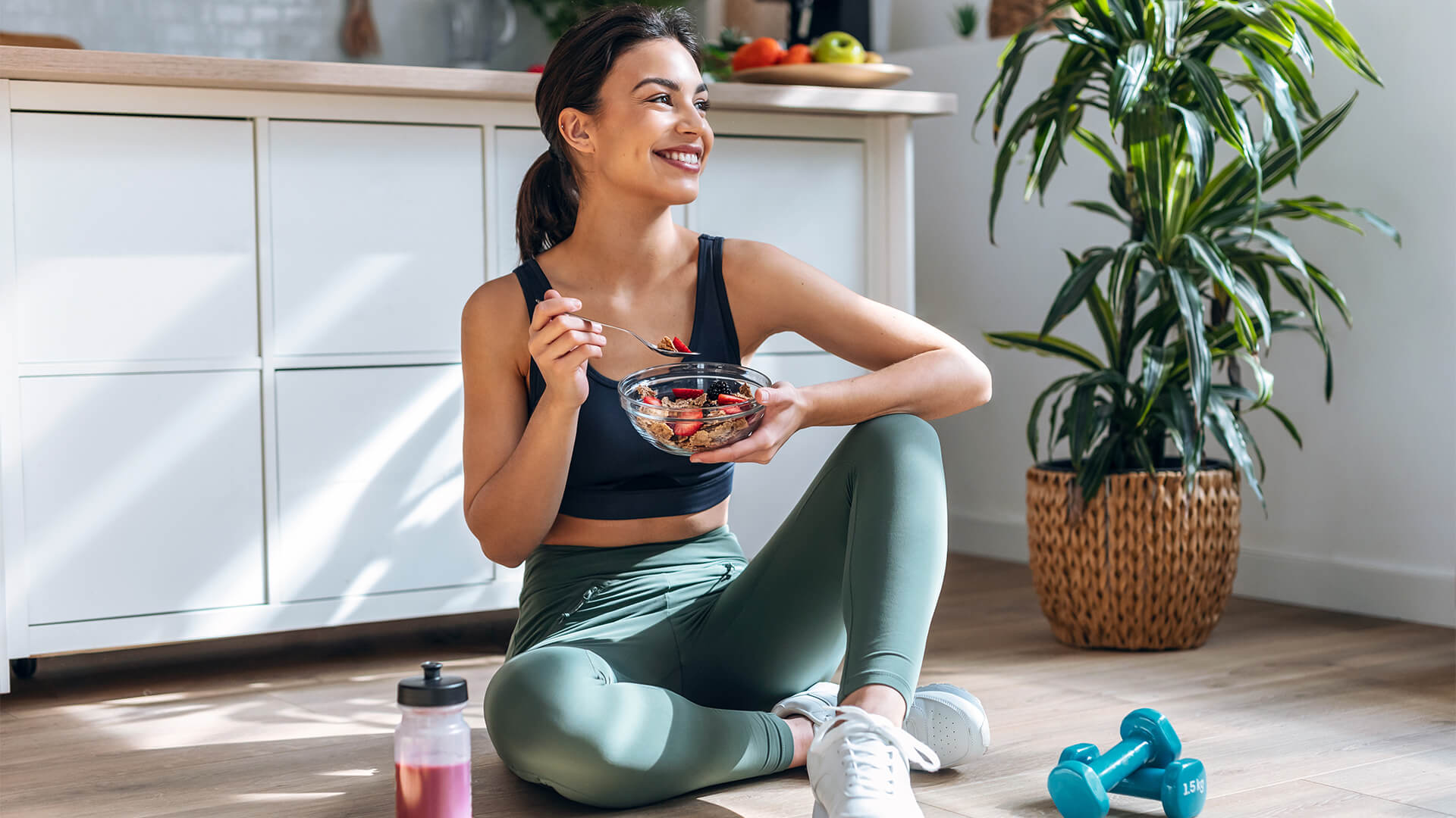 Athletic woman eating a healthy bowl of muesli with fruit sitting on floor in the kitchen at home